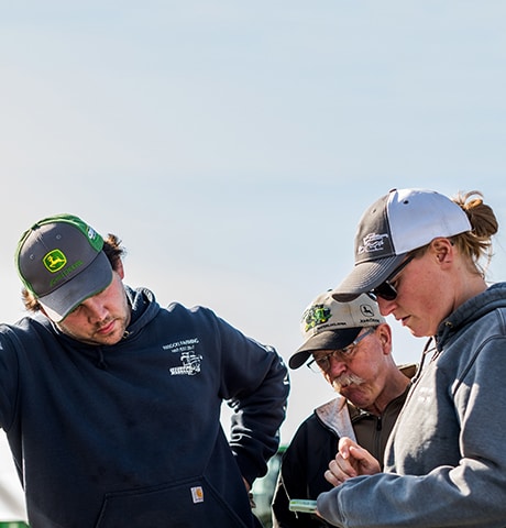 En far og hans voksne børn står ved siden af baglugen på en pickup-truck og ser på datterens mobilenhed i Alberta, Canada