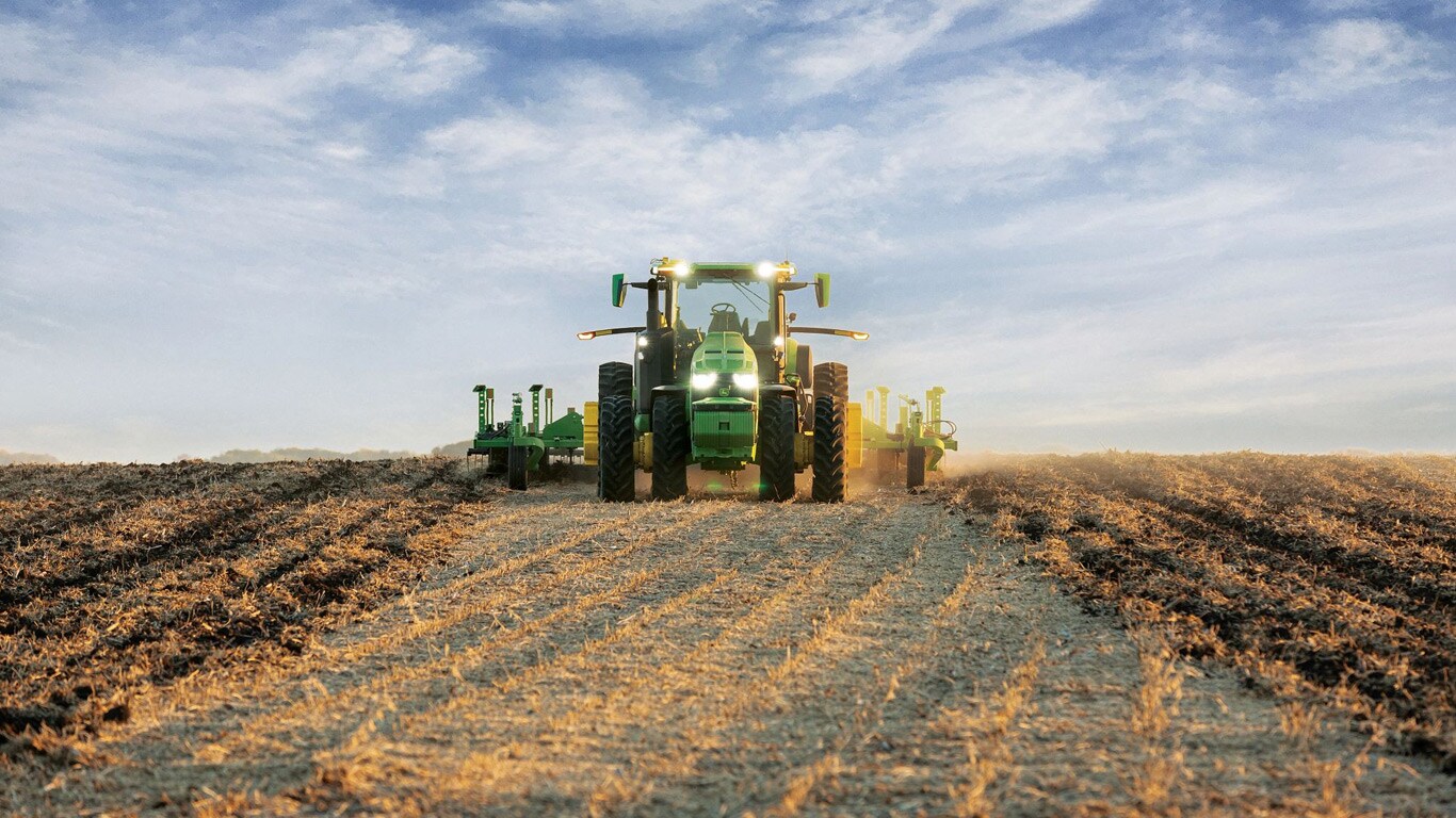 Selvkørende John Deere traktor trækker jordbearbejdningsudstyr hern over en åben mark.