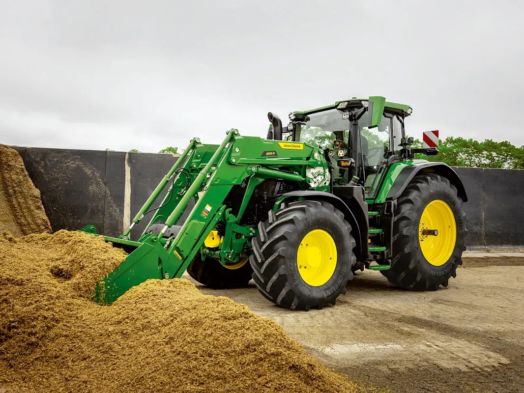 John Deere-traktor, der skubbes ned i en ensilagebunke med frontlæsserskovl i et landbrugsmiljø.