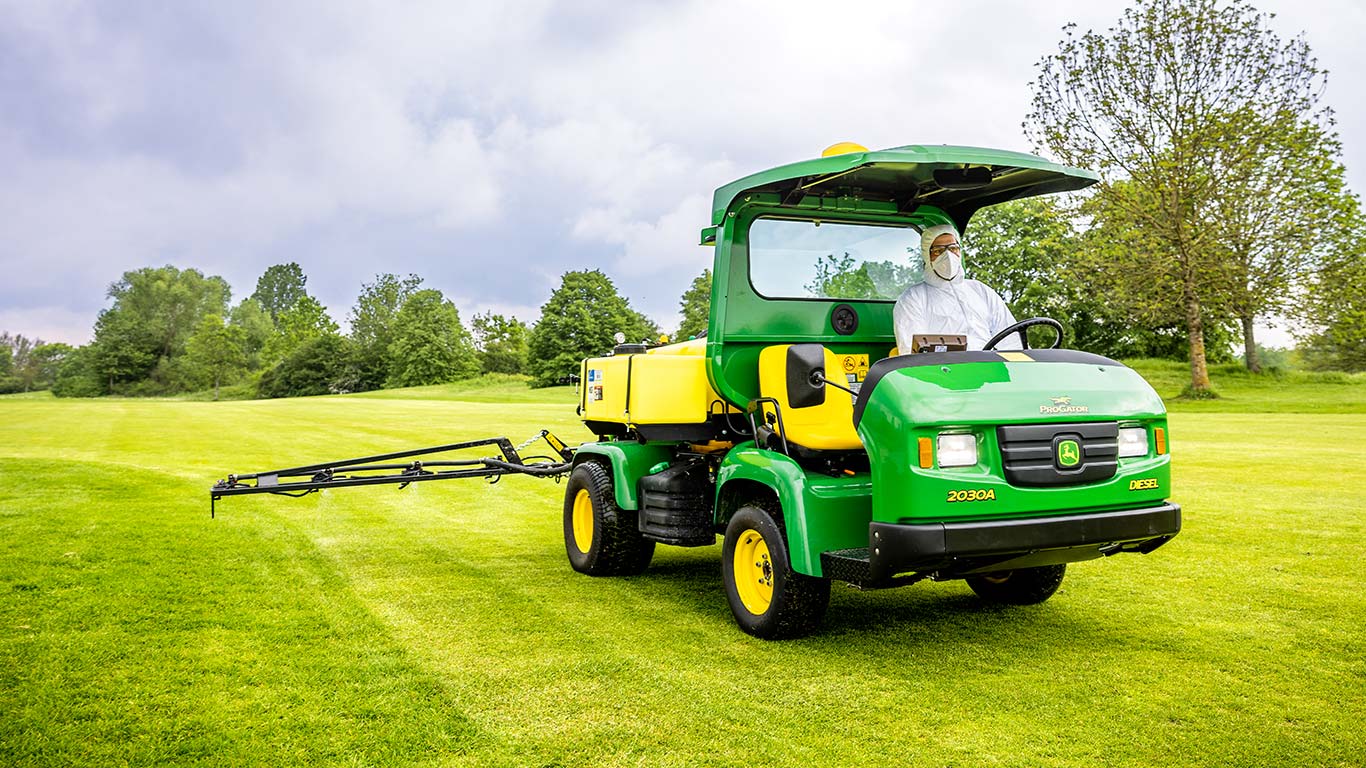 En greenkeeper i beskyttelsestøj arbejder på en golfbane med en John Deere-sprøjte.