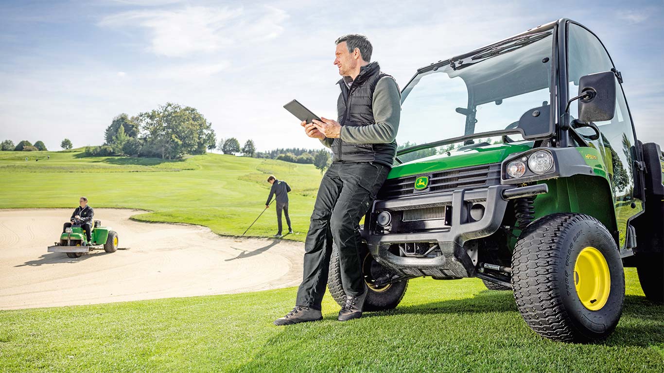 En greenkeeper kontrollerer data på sin tablet, mens 2 greenkeepere arbejder på en golfbane i baggrunden.