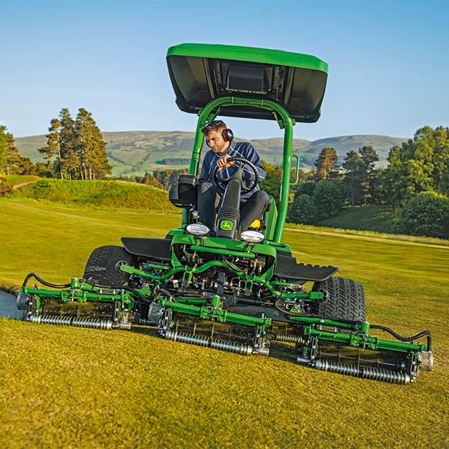 En greenkeeper arbejder på et græsområde med en John Deere-plænetraktor set forfra.