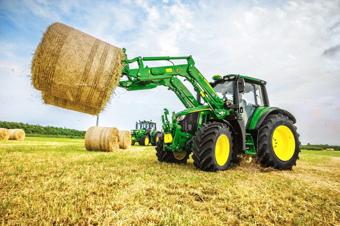 En John Deere-traktor løfter en stor rund høbal med en frontlæsser på en høstet mark under en blå himmel