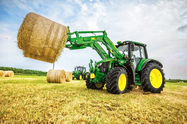 En John Deere-traktor løfter en stor rund høbal med en frontlæsser på en høstet mark under en blå himmel