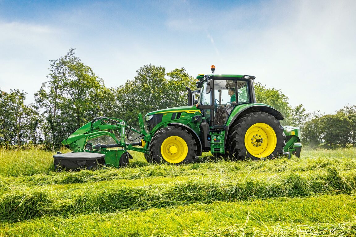 En John Deere-traktor, der slår græs med en frontmonteret slåmaskine på en grøn eng tæt på træerne