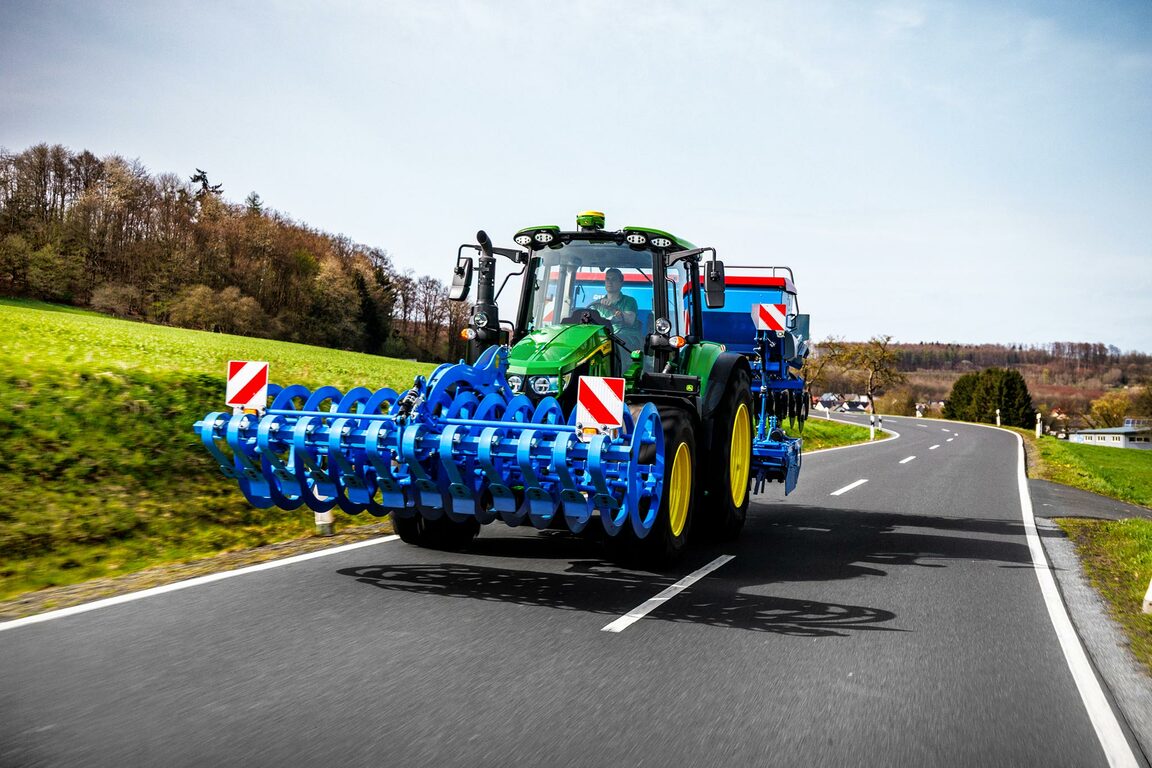 En John Deere traktor, der transporterer en bred blå kultivator på en landevej med sikkerhedsmarkeringer