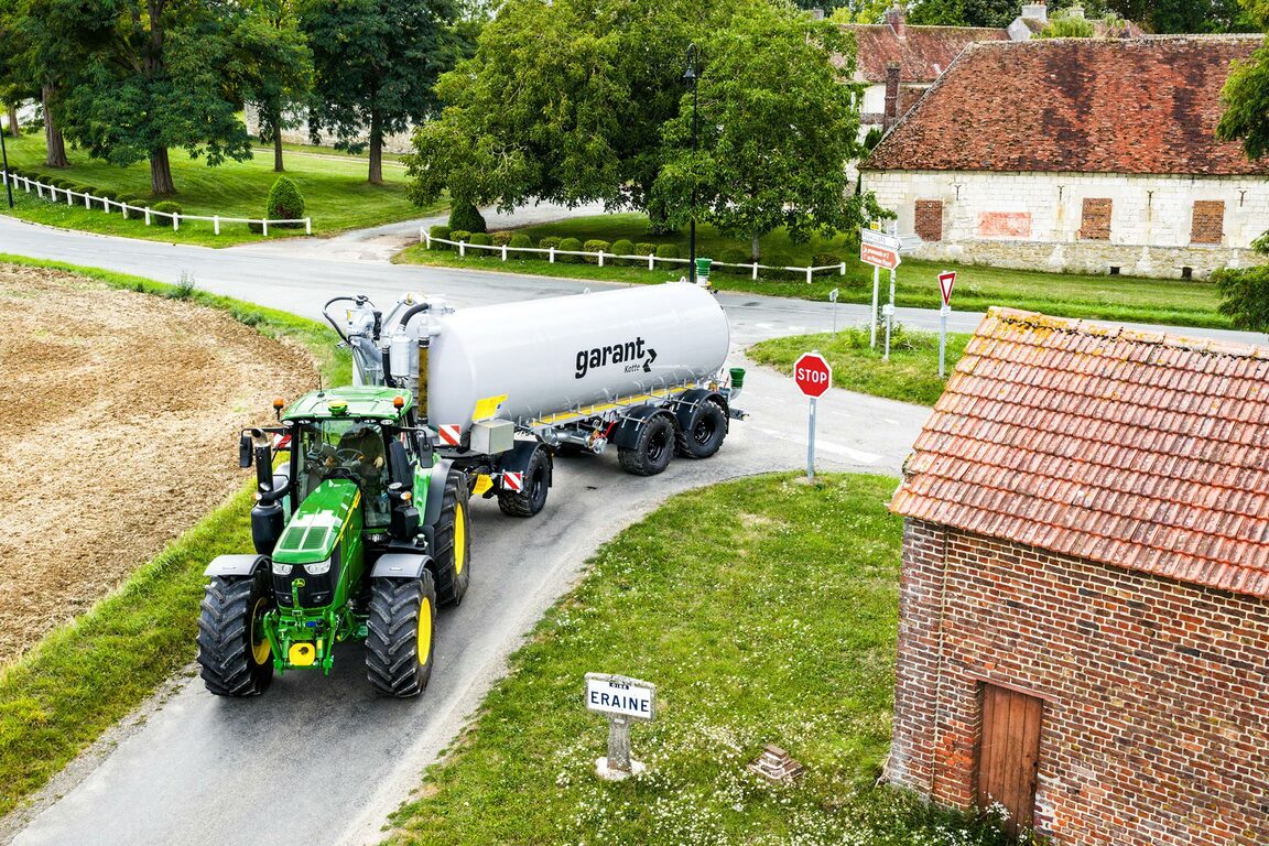 En John Deere-traktor, der trækker en tankvogn til flydende gylle på en landevej i nærheden af landbrugsbygninger