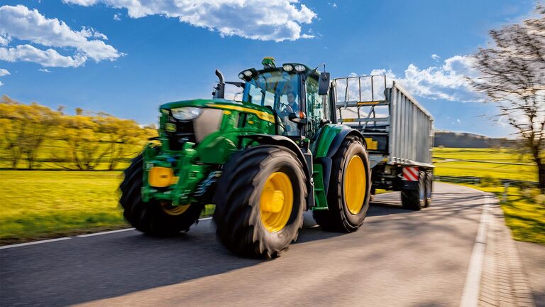 En John Deere-traktor trækker en landbrugsvogn på en landevej gennem grønne marker under en blå himmel med spredte skyer.