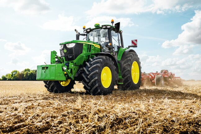 John Deere traktor, der trækker et rødt jordbearbejdningsredskab hen over en støvet høstet mark
