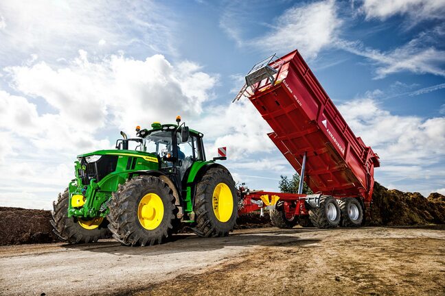 En John Deere-traktor læsser materiale af en rød tippelad på en landbrugsgård