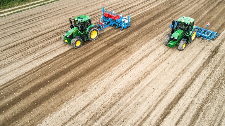 To John Deere 6M traktorer p&aring; en mark med bar jord. Den ene tr&aelig;kker en Monsomen planter.  Den anden tr&aelig;kker Lemken s&aring;bedskombination