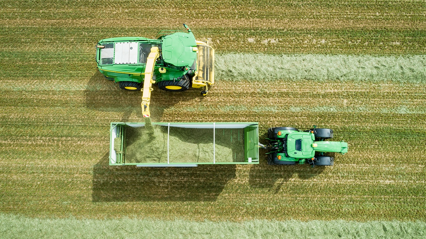 Luftfoto af John Deere 8000 serie selvk&oslash;rende finsnitter med gr&aelig;sopsamling til vogn trukket af John Deere traktor