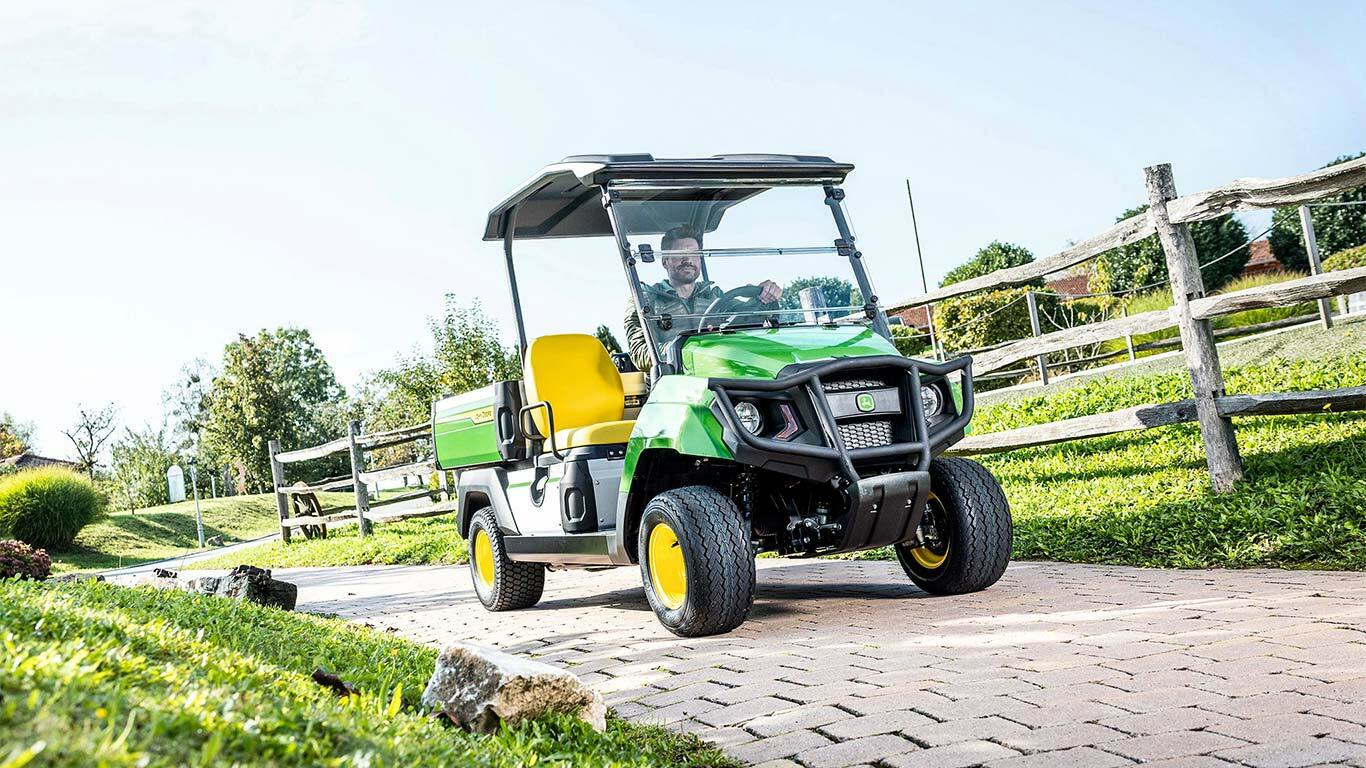 Grøn John Deere Gator GS transporter, der kører på en asfalteret sti gennem et anlagt område med græs og træhegn.