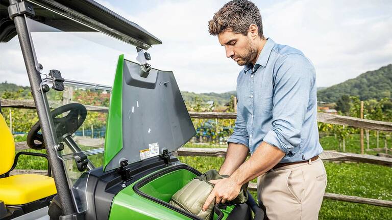 Person, der anbringer en polstret pose i opbevaringsrummet foran på en grøn John Deere Gator GS, som er parkeret udendørs i nærheden af et træhegn og en vingård.