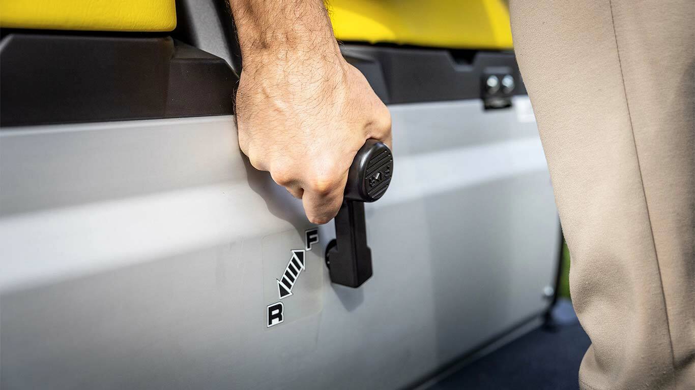 Nærbillede af en hånd, der betjener gearstangen på en John Deere Gator GS med gule sæder, hvor positionerne for frem og bak er vist.