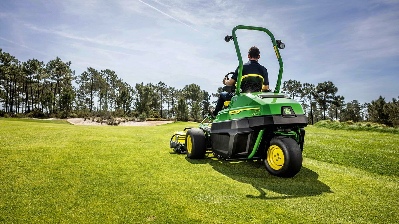En person kører en grøn elektrisk John Deere 2775 E-Cut™ greenklipper over en velholdt golfbane med træer og en delvist overskyet himmel i baggrunden.