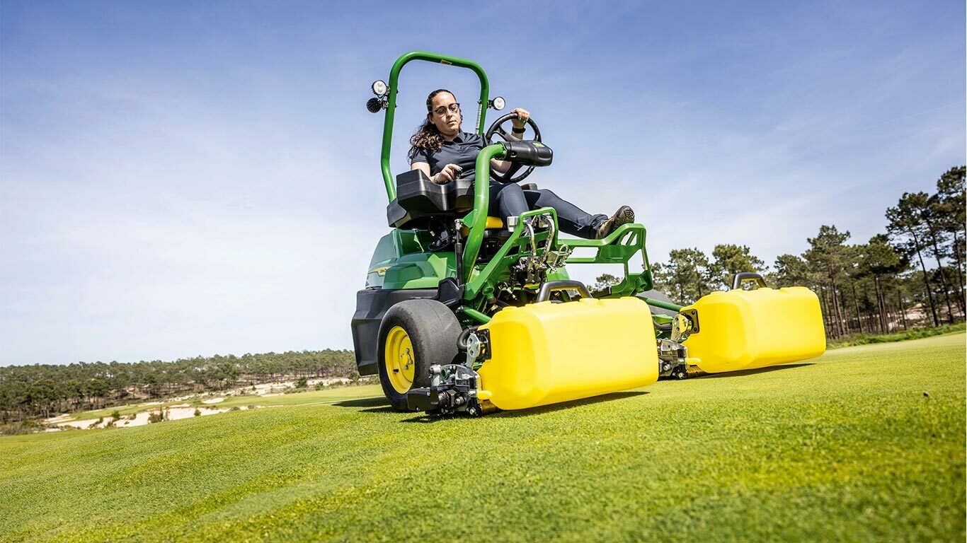 En person kører en grøn og gul elektrisk John Deere 2775 E-Cut™ plæneklipper med frontruller dækket af et gult hus på en golfgreen med træer og en klar blå himmel i baggrunden.