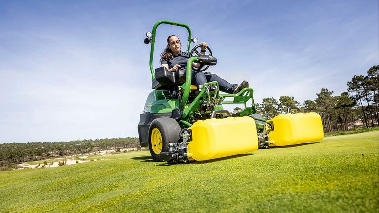 En person kører en grøn og gul elektrisk John Deere 2775 E-Cut™ plæneklipper med frontruller dækket af et gult hus på en golfgreen med træer og en klar blå himmel i baggrunden.