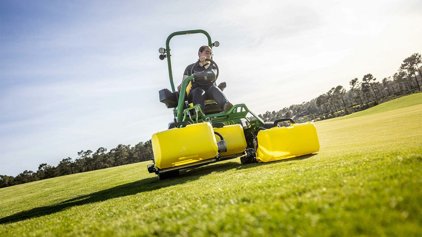 En person kører en grøn og gul elektrisk John Deere 2775 E-Cut™ plæneklipper med tre store frontruller dækket af gule huse på en golfgreen med træer og en delvist overskyet himmel i baggrunden.