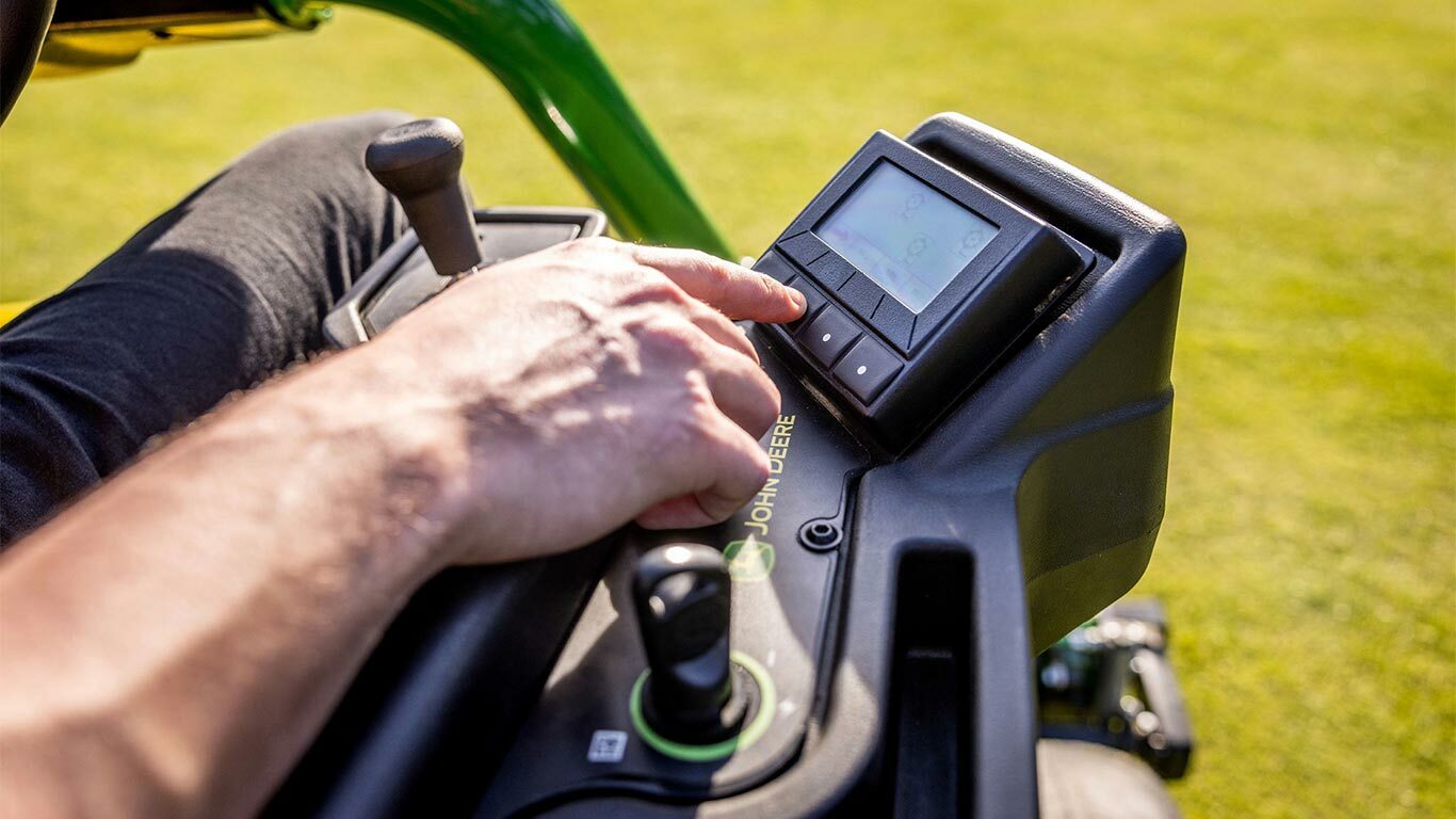 Nærbillede af en hånd på TechControl-panelet på en elektrisk John Deere 2775 E-Cut™ plæneklipper, hvor personen trykker på knapperne på en digital skærm med et joystick og andre synlige betjeningselementer.