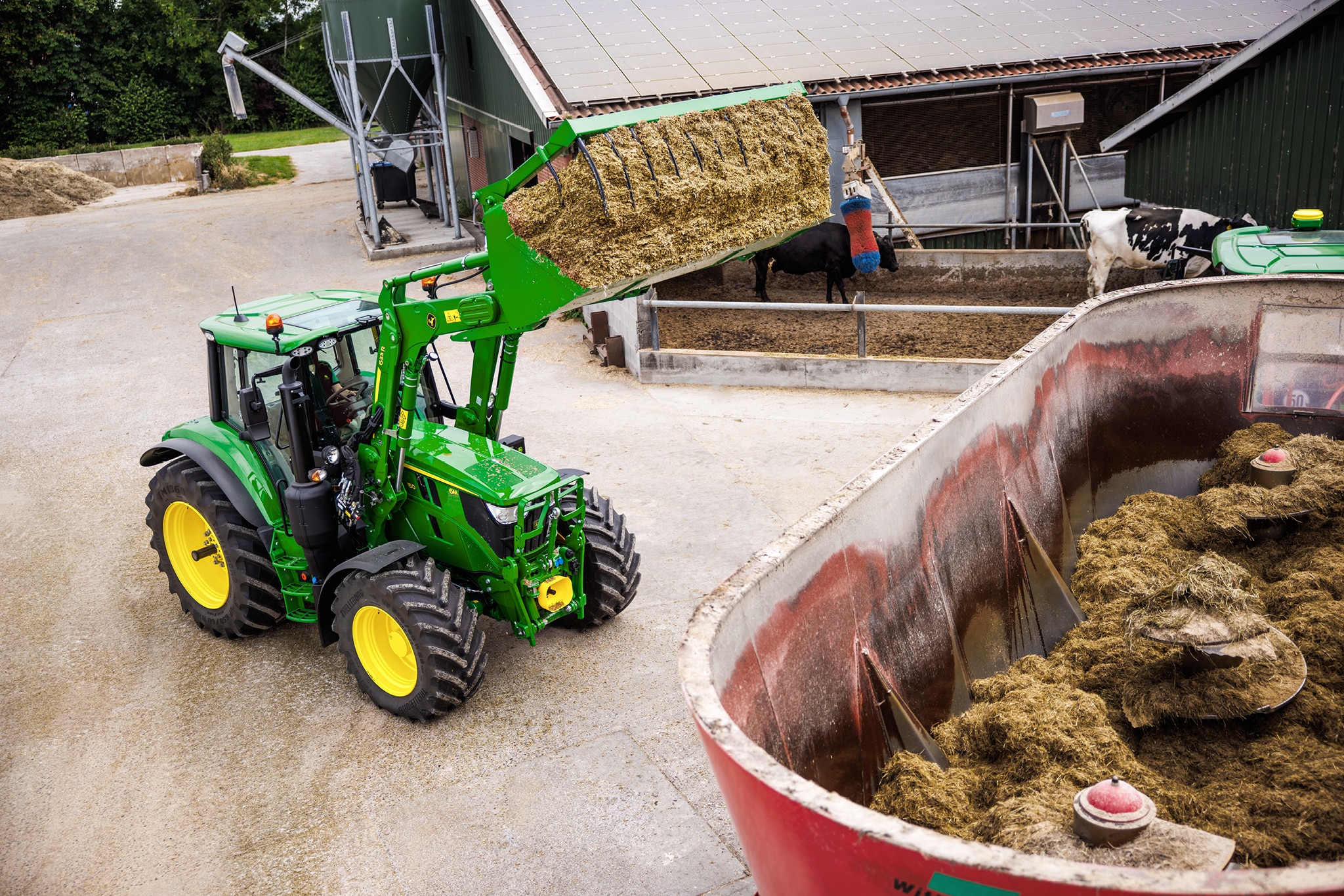 En traktor fra John Deere 6M-serien, der bruger en frontlæsser til at læsse ensilage over i en blandevogn på en gårdsplads.