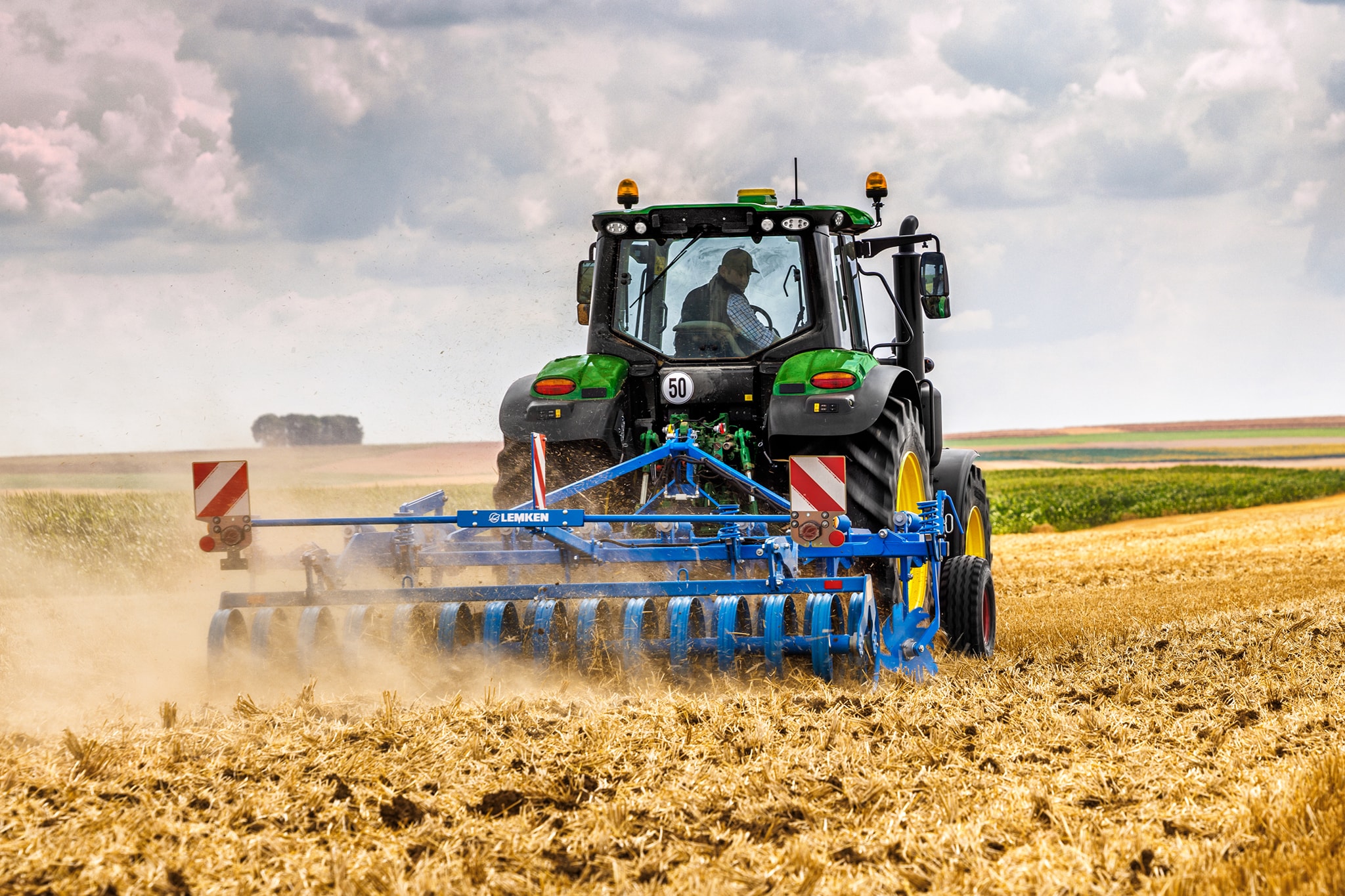 En traktor fra John Deere 6M-serien, der bearbejder en høstet mark med en skiveharve, set bagfra i støvede markforhold.