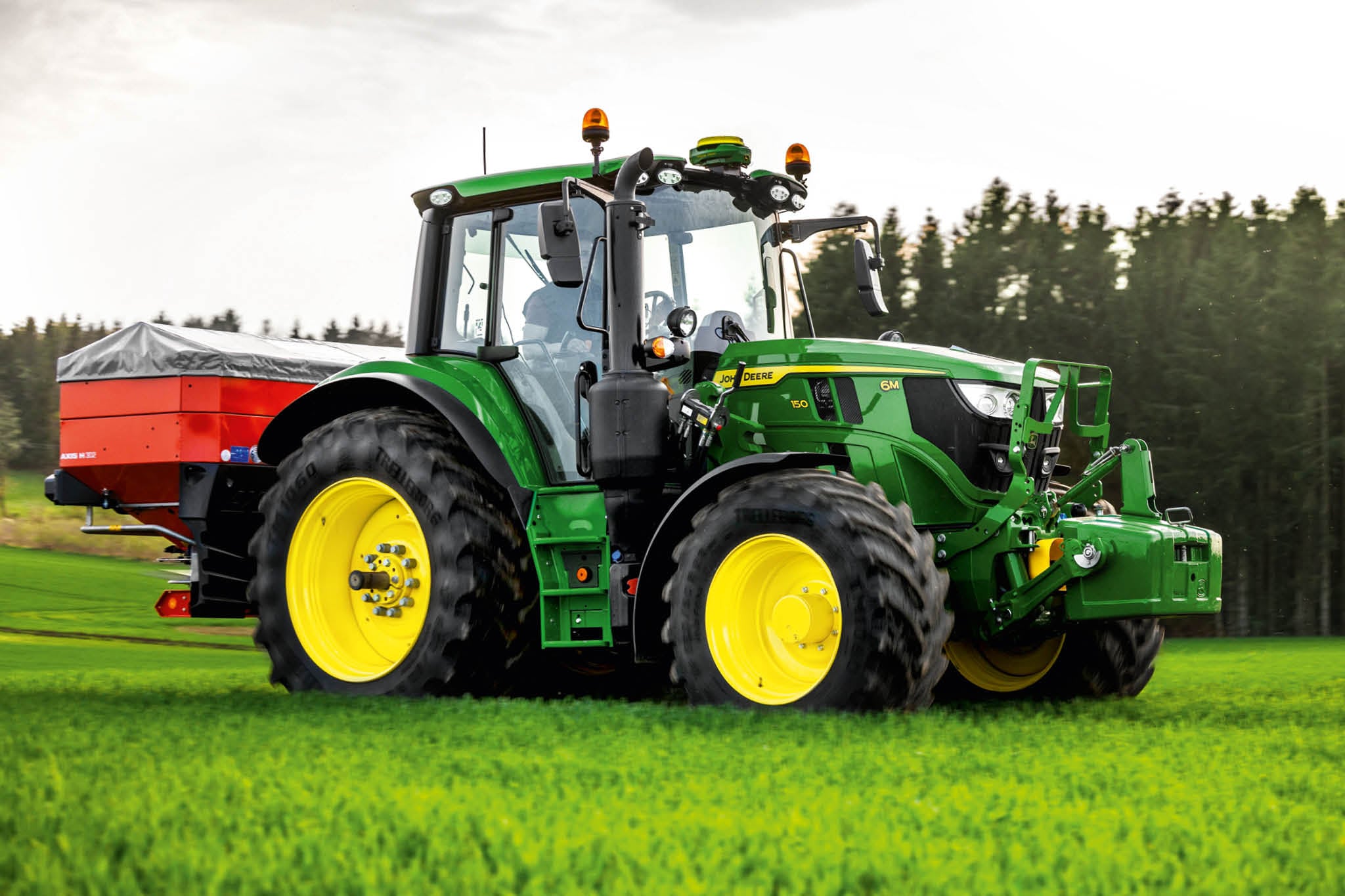 En traktor fra John Deere 6M-serien med bagmonteret spreder i aktion på en græsmark tæt på en skovkant.