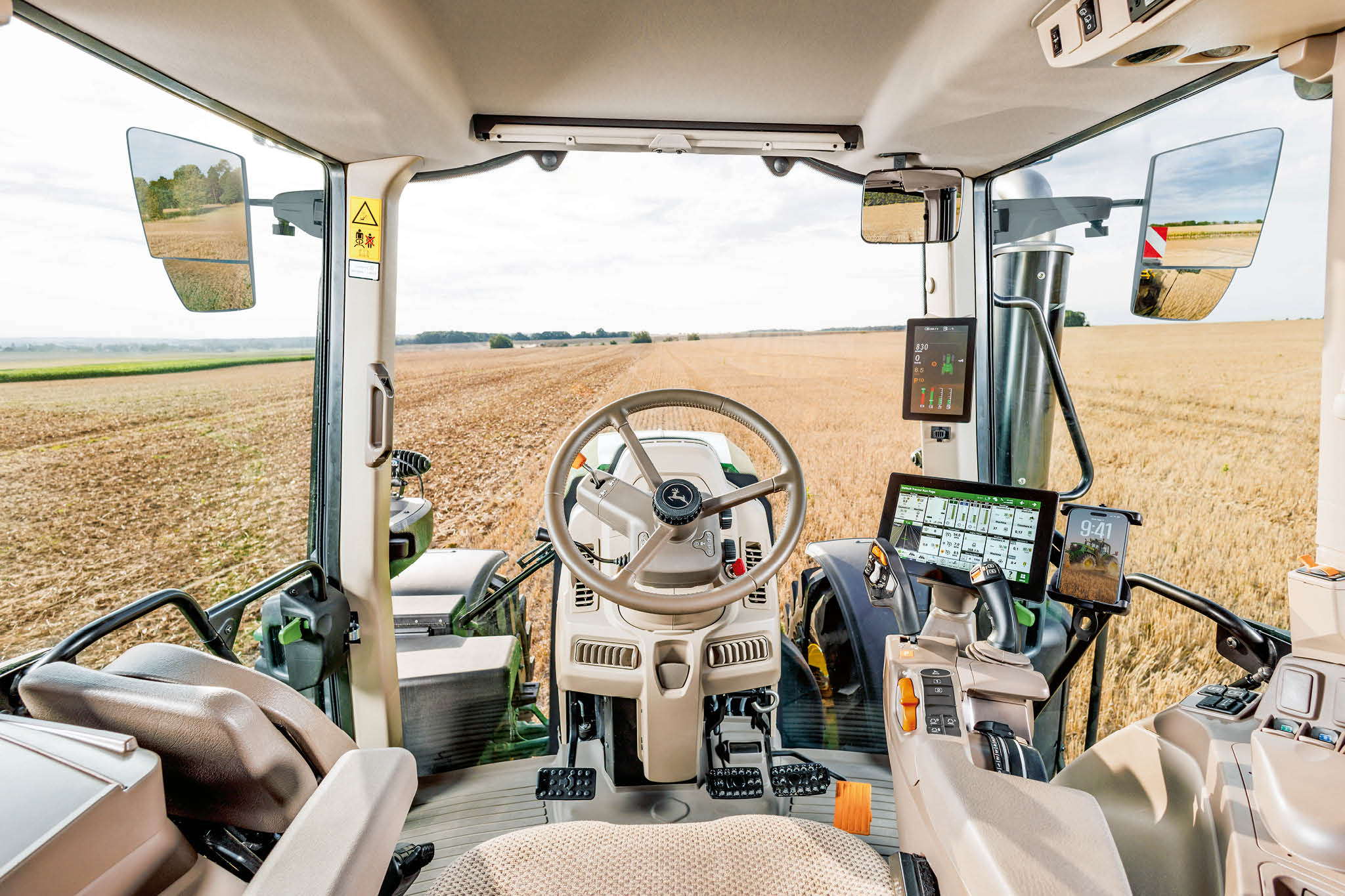 John Deere traktorkabine med rat, digitale kontrolskærme og udsigt over marken gennem forruden under arbejdet på gården.