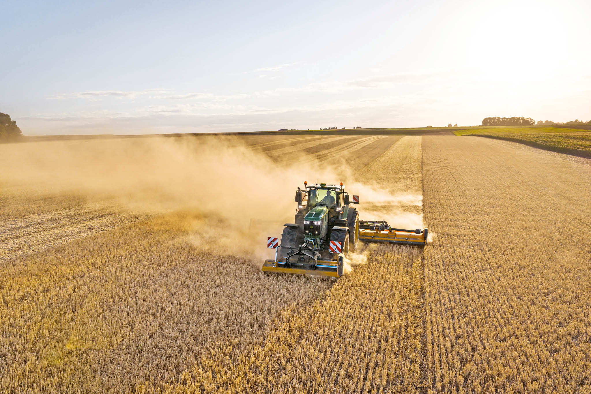 En traktor fra John Deere 6M-serien trækker en gylletankvogn ad en landevej omgivet af græs og marker.