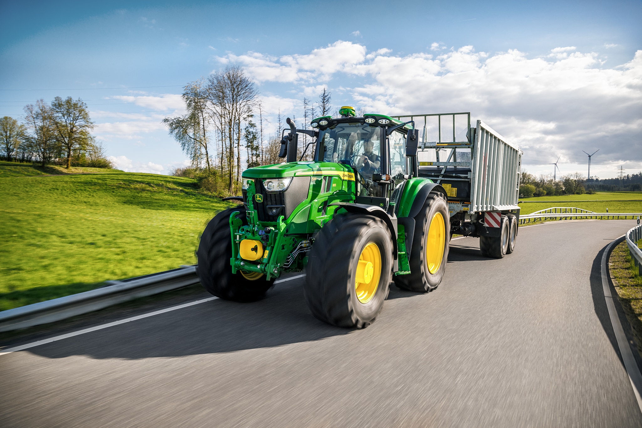 En traktor fra John Deere 6M-serien, der trækker en trailer med høje sider på en asfalteret landevej.