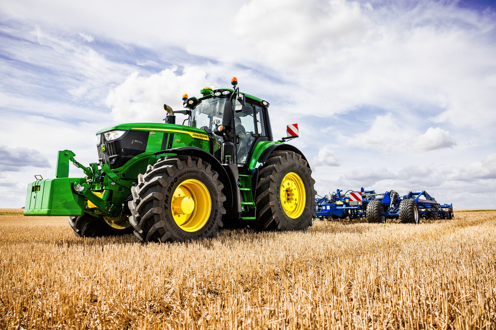 En traktor fra John Deere 6M-serien trækker et tungt jordbearbejdningsredskab hen over en stubmark under overskyet himmel.