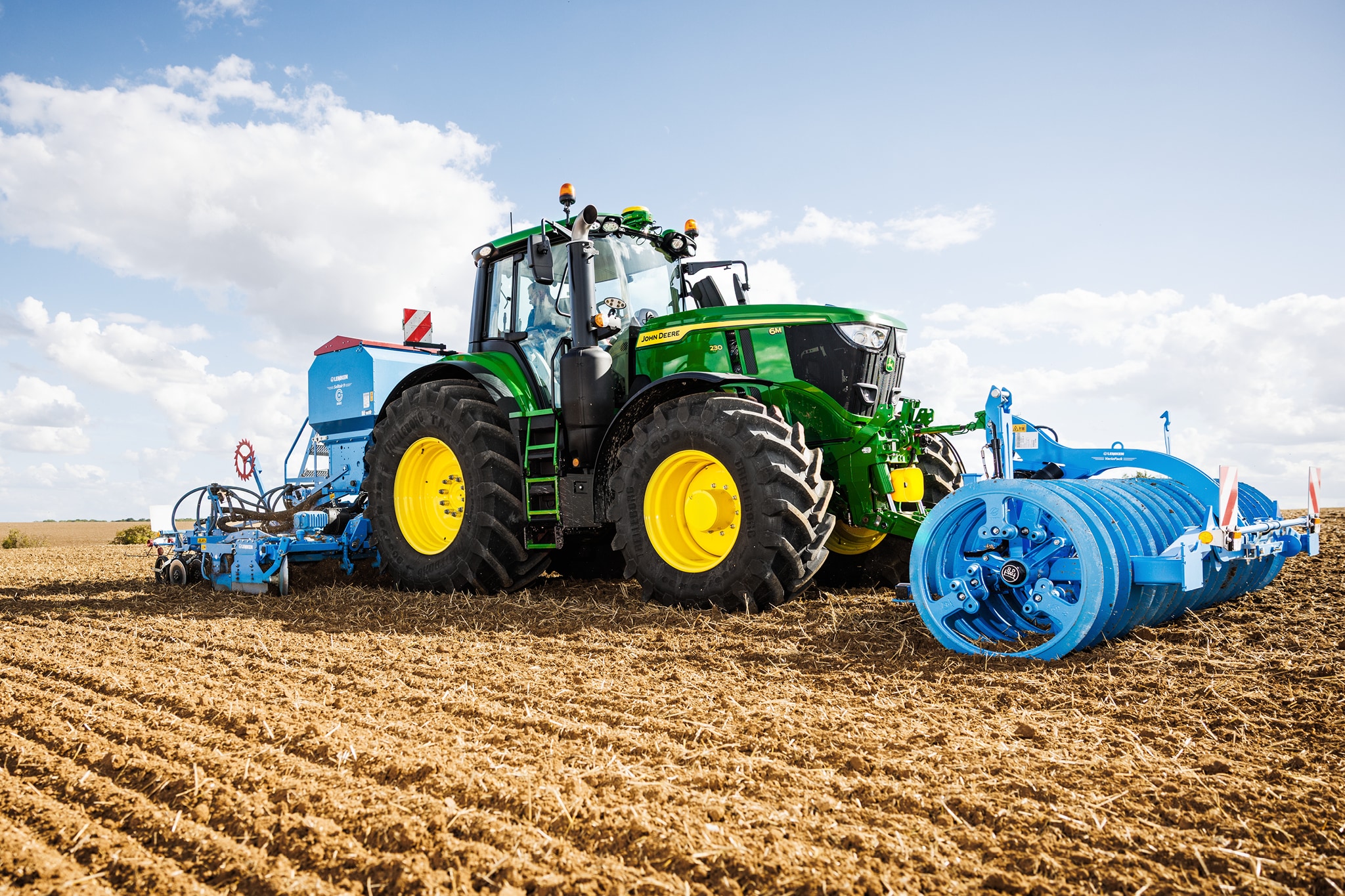 En traktor fra John Deere 6M-serien, der bearbejder en mark monteret med et stort blåt tromleredskab.
