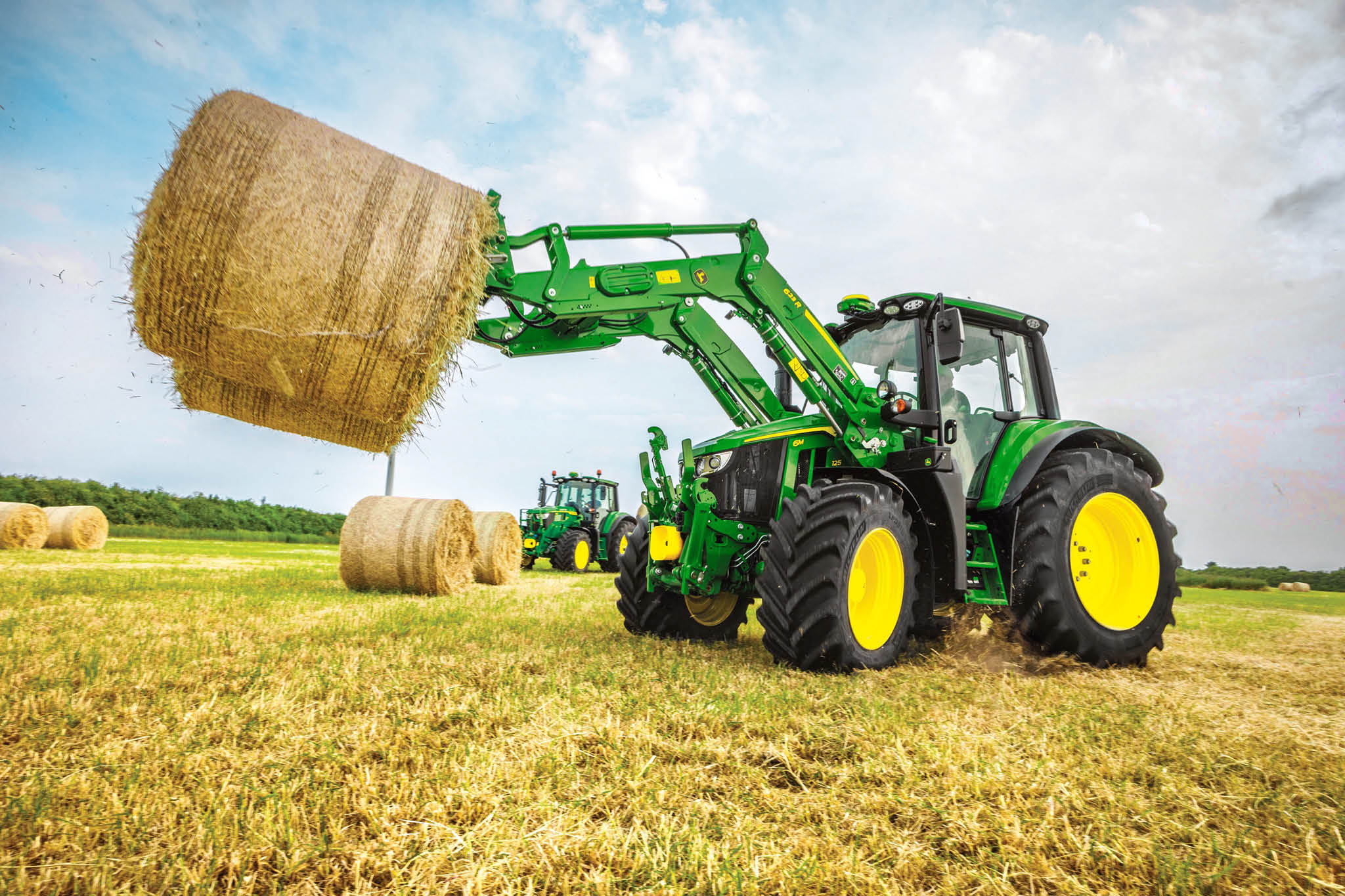 John Deere 6M serie traktor, der løfter en stor rundballe med en frontlæsser på en åben mark.