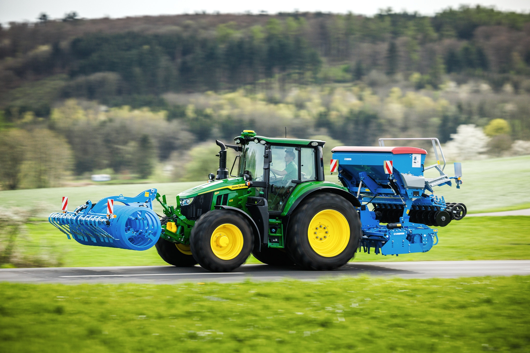 En traktor fra John Deere 6M-serien, der transporterer et frontmonteret redskab og en bagmonteret såmaskine ad en landevej.