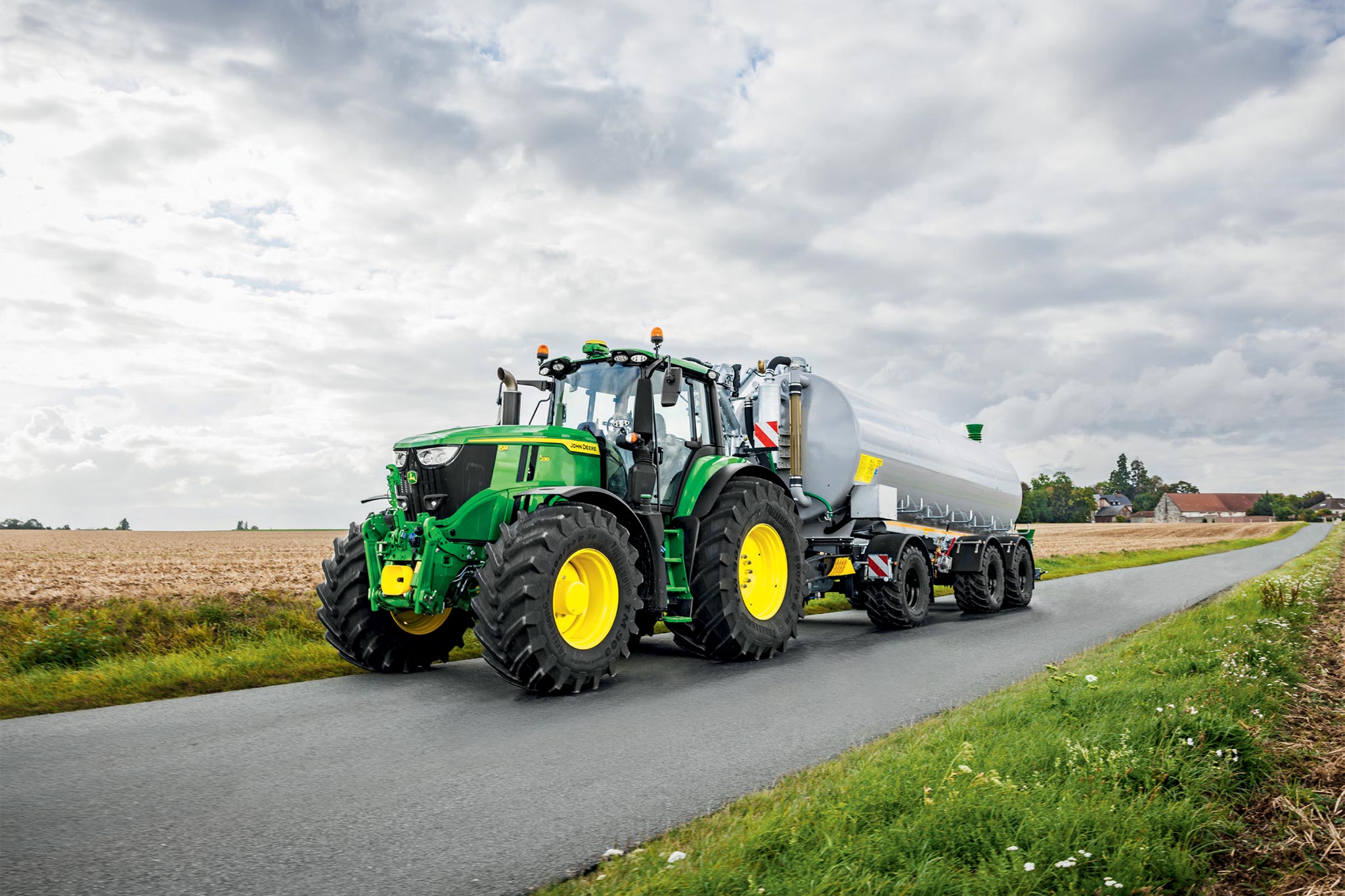 En traktor fra John Deere 6M-serien arbejder på en høstet mark med brede kultivatorer, set ovenfra med støv, der stiger op.