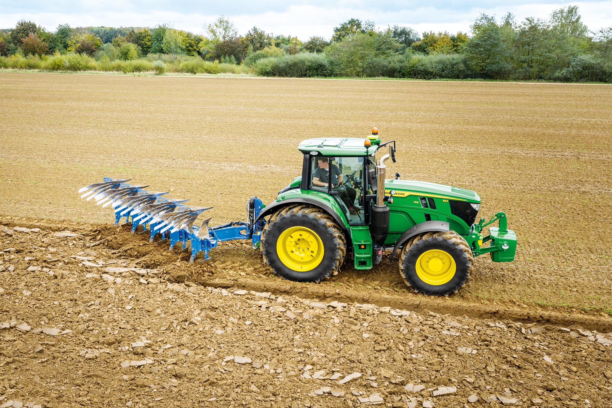 En traktor fra John Deere 6M-serien arbejder på en pløjet mark med et bagmonteret jordbearbejdning-redskab under overskyet himmel.