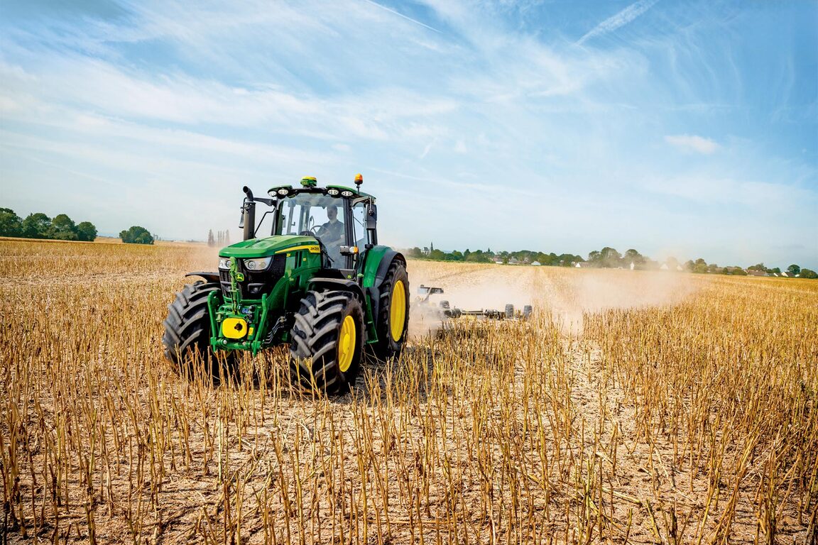 En John Deere-traktor bearbejder en høstet mark og hvirvler støv op, mens den trækker landbrugsredskaber under en klar blå himmel.