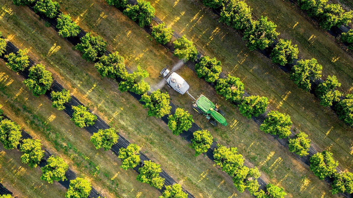 Set fra luften, grøn og gul John Deere 5015EN traktor, der bugserer en sprøjte gennem jævnt fordelte plantagerækker.