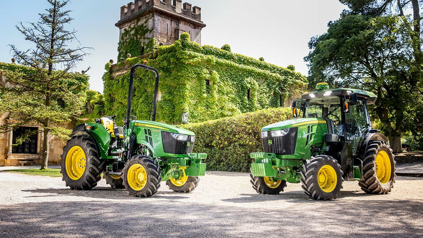 Grøn og gul John Deere 5075EN traktor, der kører gennem en plantage på en jordvej, omgivet af frugttræer og sollys.