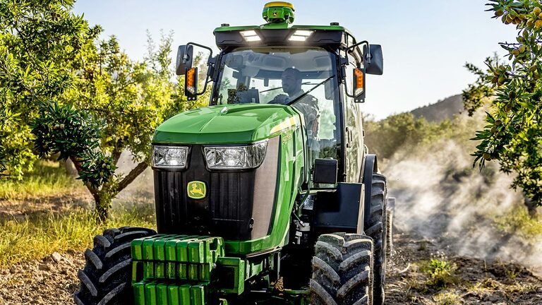 Grøn og gul John Deere 5075EN traktor, der kører gennem en plantage på en jordvej, omgivet af frugttræer og sollys.