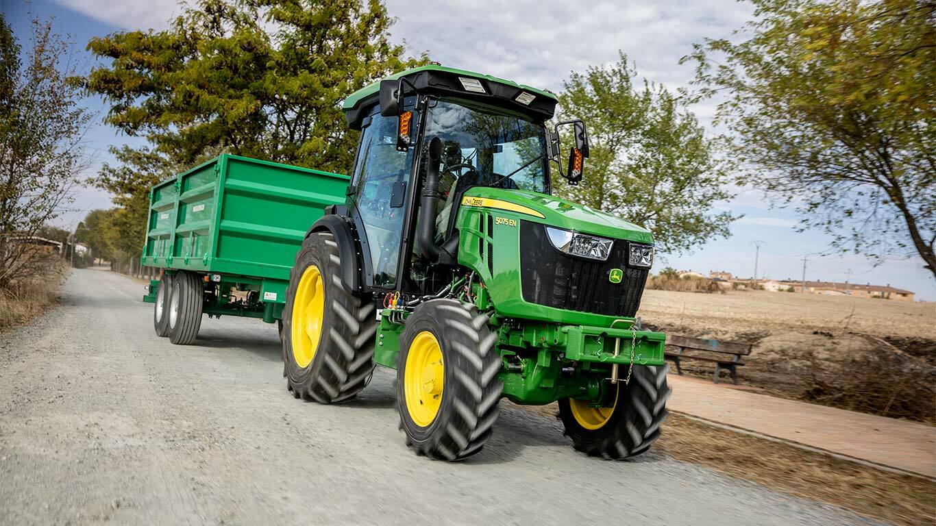 John Deere 5075EN traktor trækker en grøn trailer på en landlig grusvej med træer i baggrunden.