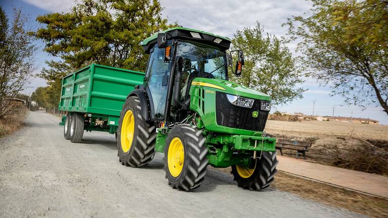 John Deere 5075EN traktor trækker en grøn trailer på en landlig grusvej med træer i baggrunden.