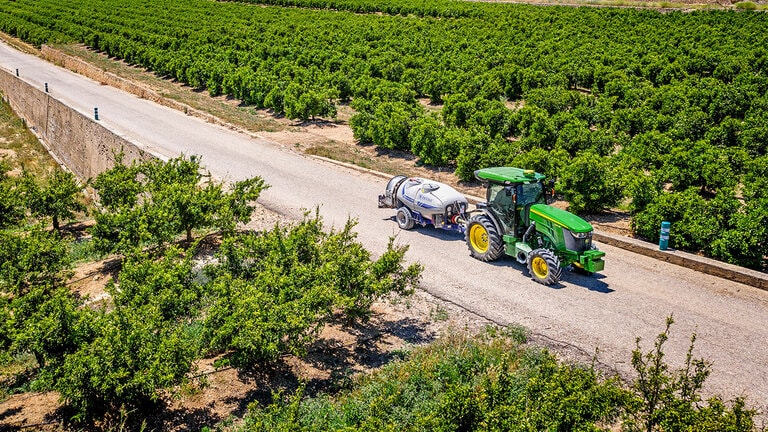 Grøn og gul John Deere 5090EN traktor, der bugserer en sprøjte på en asfalteret vej mellem store plantagerækker under en klar blå himmel.