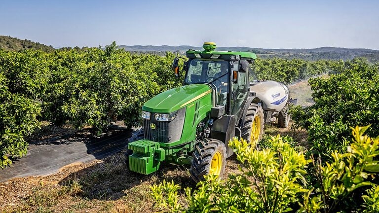 Grøn og gul John Deere 5105EN traktor, der bugserer en sprøjte i en frodig plantage med kuperede bakker i baggrunden.