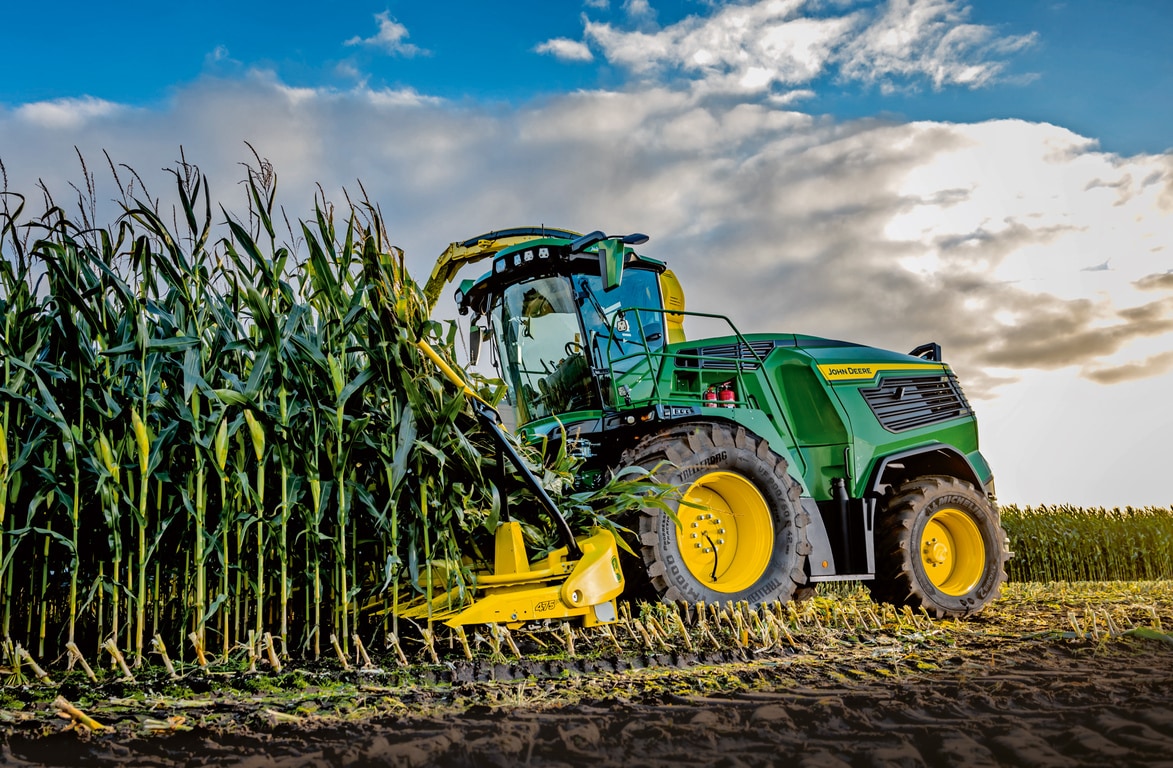 John Deere finsnitter høster majs på en mark under en blå himmel med skyer.