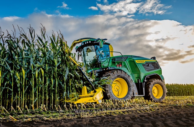 John Deere finsnitter høster majs på en mark under en blå himmel med skyer.