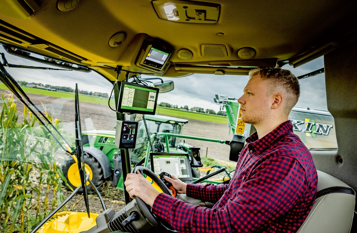Landmand, der betjener avancerede landbrugsmaskiner med flere digitale skærme i et SPFH-førerhus. Der vises moderne landbrugsteknologi på en mark.