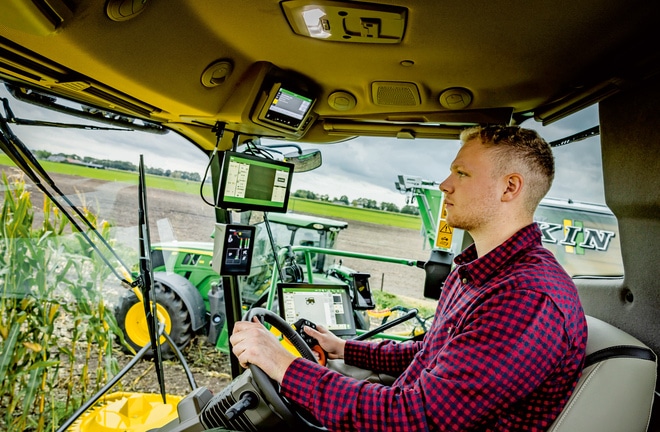 Landmand, der betjener avancerede landbrugsmaskiner med flere digitale skærme i et SPFH-førerhus. Der vises moderne landbrugsteknologi på en mark.