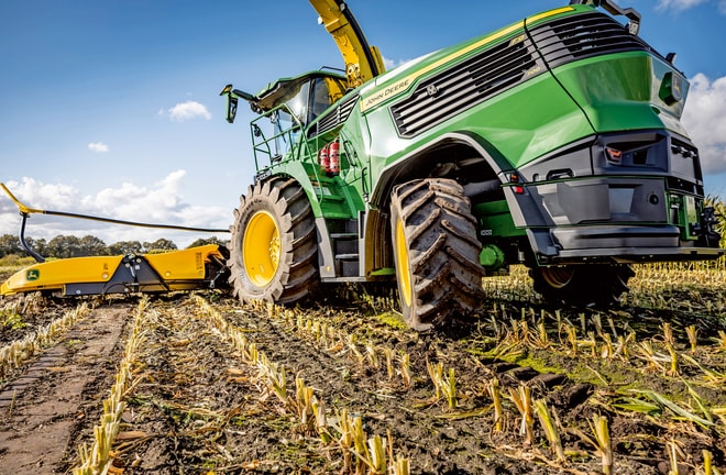 John Deere finsnitter arbejder i en høstet majsmark. Der vises maskinens store dæk og skæremekanisme mod en klar blå himmel.