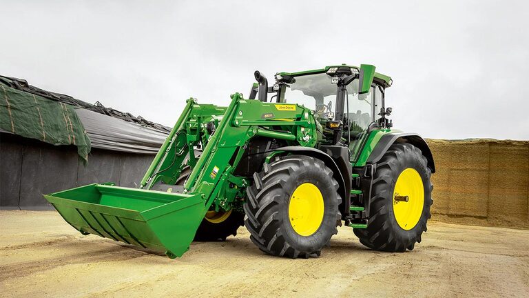 John Deere traktor med frontlæsser parkeret på en gårdsplads tæt ved opbevaring af ensilage.